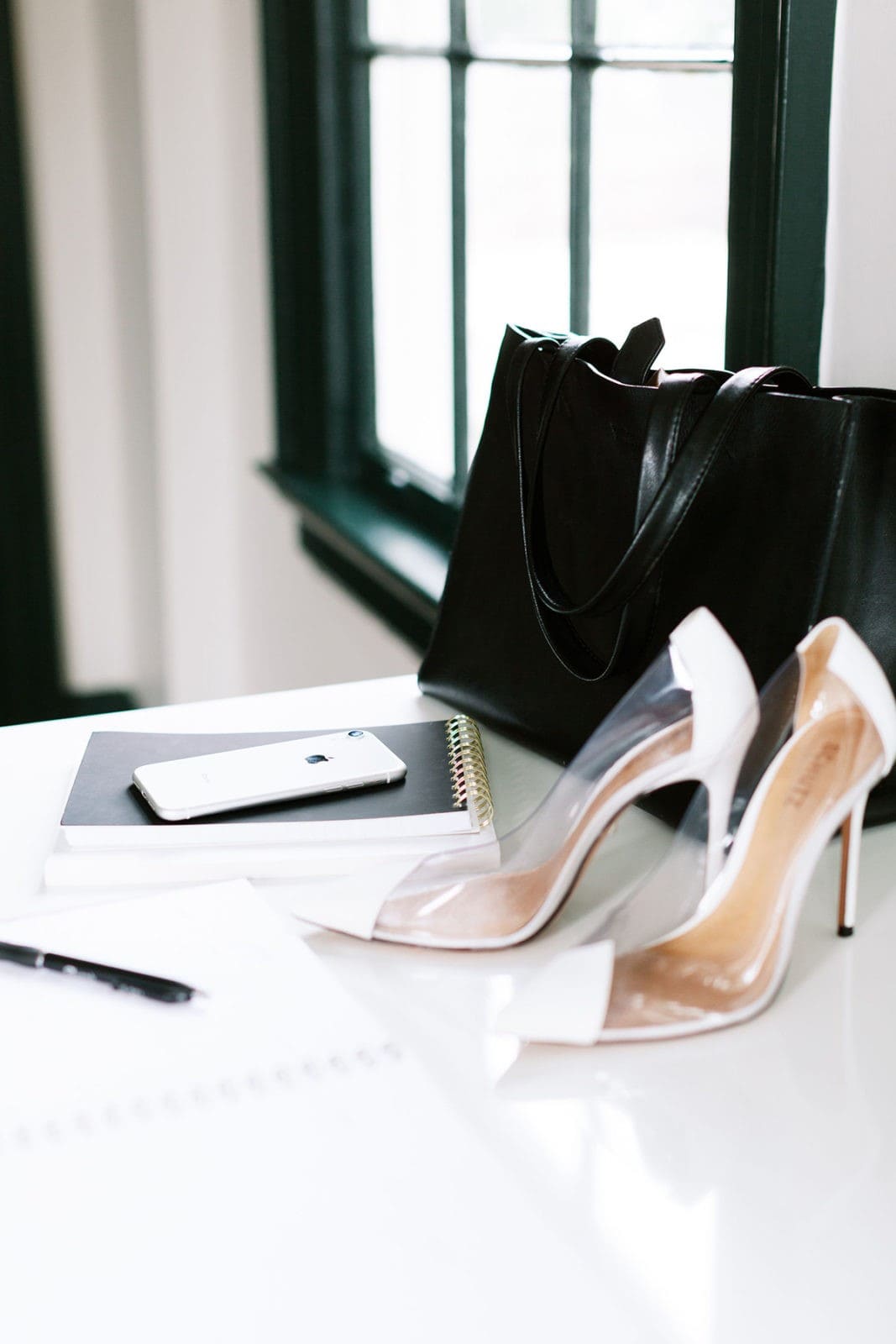 Heels, black purse, and iphone on white desk.